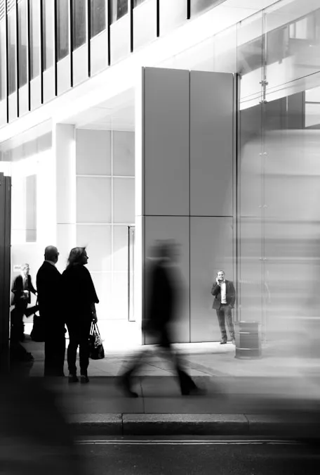 Black and white photo of people standing and walking outside a modern building with large glass windows and geometric panels.